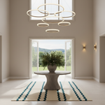 A modern multi-ring chandelier with a warm glowing light hangs in a bright, high-ceilinged entryway. The room features a stone pedestal table with a potted green plant, a cream and teal patterned rug, and large windows looking out to a grassy field.