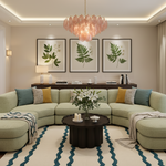 A modern living room featuring a curved light green sectional sofa with yellow and teal accent pillows, a round black coffee table, a pink glass chandelier, and three framed botanical prints on the beige wall.