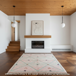 A modern living room interior with a light pink and mint geometric patterned area rug on a dark wood herringbone floor, positioned in front of a white wall with a fireplace and wooden mantel.