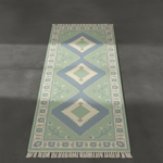 A rectangular area rug with a traditional geometric pattern in sage green, light blue, and cream, featuring white fringe on the ends, shown from a top-down perspective on a dark gray floor.