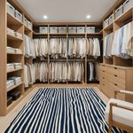 A spacious, well-organized walk-in closet with light wood cabinetry, shelves, drawers, and hanging garments. A large navy blue and cream striped rug covers the floor, and a portion of a matching upholstered chair is visible in the lower right corner.