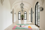 A colorful abstract-patterned area rug is displayed on a light-colored stone hallway with arched windows, black metal lanterns, and wall sconces.