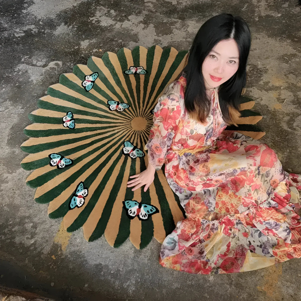 Woman in a floral kimono sitting next to a large decorative fan with butterfly designs on a textured floor.