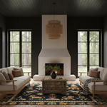 A styled living room interior with two cream sofas, a wooden coffee table, a large patterned rug, a white fireplace, and a tiered woven pendant light. Large windows with dark trim show a forest view outside.