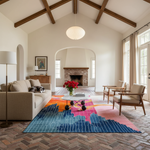 A large, vibrant abstract-patterned area rug in blue, pink, orange, and white is styled in a spacious, high-ceilinged living room with a brick fireplace, wooden ceiling beams, and mid-century modern furniture.