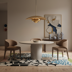 A modern dining room scene featuring a round white stone dining table, four upholstered dining chairs in a warm taupe-brown fabric, a large brass pendant light, and a bold black, white, and blue patterned area rug on a light wood floor.