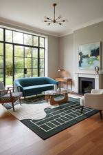 A styled living room interior featuring a teal blue velvet loveseat as the primary furniture piece, set on a geometric patterned rug with a glass-top coffee table, wood flooring, a fireplace, and a large window view.