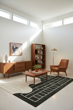 A modern mid-century styled living room featuring a warm brown leather armchair, a matching wooden sideboard, a wooden bookshelf, and a black and beige patterned area rug on a speckled floor.