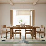 A minimalist dining room scene featuring a light oak wood dining table and six matching wooden chairs, styled with neutral ceramic vases and a green plant, set on a patterned abstract area rug.