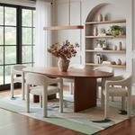 A modern dining room scene featuring a round wooden dining table with four upholstered cream chairs, styled on a patterned rug with a vase of foliage in the center. The background shows a white wall with built-in shelves, a window with black frames, and a hanging light fixture.