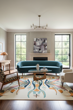 A styled living room featuring a teal velvet sofa centered in front of a fireplace, with a large abstract painting above it. The room includes a patterned area rug, a glass-top coffee table, a wooden chair, a white armchair, and tall black-framed windows looking out onto greenery.