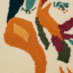 Close-up of a textured area rug with a colorful abstract pattern on a cream background, featuring prominent orange, mustard yellow, dark green, burgundy, and teal sections.