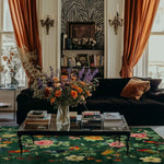 Elegant living room featuring the Emerald Oasis Hand Tufted Area Rug. The vibrant wool rug with floral patterns enhances the space, complemented by a plush black sofa and a coffee table adorned with books and a colorful flower arrangement. Perfect for living room and dining room settings.