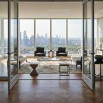 A luxury modern living room with floor-to-ceiling windows overlooking a city skyline, furnished with two black armchairs, glass tables, a floral area rug, and a geometric side table, viewed from inside through open glass doors.