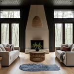 A modern living room with a large central fireplace, dark wood trim, and tall windows looking out onto a wooded forest. Two cream-colored sofas face each other across a round woven coffee table, with a blue patterned rug on the floor in front.