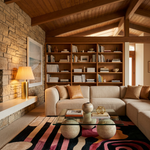 A modern living room featuring a beige sectional sofa, a large wooden bookshelf filled with books, a stone wall, a glass coffee table, and a colorful abstract patterned rug.