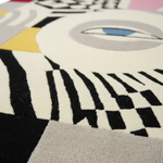 Close-up of a textured area rug featuring a modern abstract design with black, cream, gray, yellow, pink, and blue geometric shapes and a stylized eye motif.