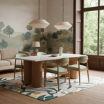 A styled dining room scene featuring a rectangular white dining table with a thick round wooden pedestal base, surrounded by six light green upholstered wooden chairs. The set is arranged on a colorful abstract patterned area rug in a bright room with a beige sofa, a tree-patterned wallpaper, and a large window looking out onto green trees.