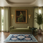 A styled interior scene with a large framed classical portrait on a textured green wall above a green chaise lounge. The room includes cream curtains, two gold wall sconces, a potted palm, dark green nesting side tables, and a blue floral area rug on a wood floor.