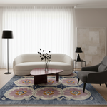 A styled living room scene featuring a cream curved sofa, a dark gray armchair, a low wooden coffee table, a large blue and pink floral area rug, a black floor lamp, and a black table lamp against a beige wall with sheer white curtains.