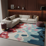 A modern abstract area rug with a multicolor design in shades of blue, pink, red, and beige, styled in a contemporary living room setting with a cream sectional sofa and dark wood paneling.