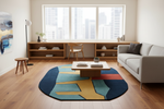 A modern living room scene featuring a prominent round area rug with a bold geometric pattern in navy blue, teal, mustard yellow, and red. The rug is styled on a light wood floor in front of a low wooden coffee table and a light gray sofa, with a large window and city view in the background.