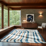 A large rectangular area rug with wavy blue, beige, and brown stripes on a cream base, styled in a modern mid-century modern living room with a wood credenza, stone wall, and large windows.