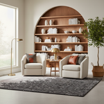 A styled living room scene with two cream upholstered armchairs facing a small wooden side table and gold desk lamp, set in front of a large built-in wooden bookshelf. A patterned area rug covers the floor, with a tall potted tree on the right and a floor lamp on the left. No people or animals are present.