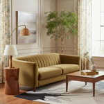 A modern olive green velvet loveseat sofa in a beautifully styled living room with a wooden coffee table, a floor lamp, a potted plant, and floral curtains.