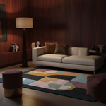 A modern living room scene featuring a large abstract area rug with geometric shapes in teal, pink, mustard, and cream, styled under a cream sofa and a dark wood-paneled wall.