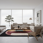 A modern living room featuring a light beige sectional sofa, a matching armchair, a low wooden coffee table, and a large abstract area rug with black, burgundy, and beige sections. The room is styled with a minimalist abstract wall art print, a floor lamp, and a large window looking out onto a tree.