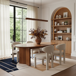 A modern dining room scene featuring a light oak wood oval table with a thick pedestal base, surrounded by four cream upholstered chairs. A large brown ceramic vase filled with autumn foliage sits on the table. The room has white walls, a built-in wooden arched shelving unit on the right, and a large window with sheer curtains on the left.