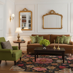 A styled living room interior featuring a brown sofa with green patterned pillows, a dark wood coffee table, a green armchair, a floral area rug, wall sconces, and two ornate gold-framed mirrors on white paneled walls.