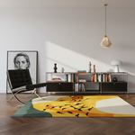 A modern living room scene featuring a large abstract area rug with yellow, orange, green, and white sections and black fish-like motifs. The rug is placed on a herringbone wood floor next to a black leather lounge chair and a low metal bookcase with books and decor.