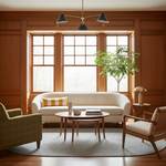 A styled living room interior with a cream curved sofa, round wooden coffee table, two accent chairs, wood-paneled walls, large windows, and a potted tree.