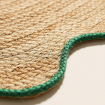 Close-up of a woven natural fiber product, likely a rug or mat, showing a textured tan jute surface with a contrasting green braided edge on a light neutral background.