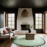 A styled living room interior with a white sofa, woven pendant light, round green rug, bamboo coffee table, dark wood-paneled walls, and large multi-pane windows.