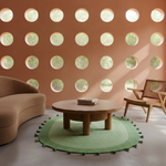 A modern living room scene with a round wooden coffee table centered on a green circular rug, a curved tan sofa on the left, and a wooden armchair on the right. The background features a terracotta-colored wall with evenly spaced circular cutouts showing outdoor greenery.