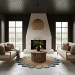 A modern living room with two light wood armchairs, a round wooden coffee table, and a large circular woven rug in front of a white fireplace and dark wood-paneled walls.