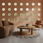 A styled living room corner featuring a curved brown sofa, a matching armchair, a round wooden coffee table, and a scalloped blue and beige rug in front of a textured terracotta wall with circular cutouts.