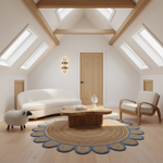 A bright, modern attic living room with light wood ceiling beams and skylights, featuring a cream curved sofa, a wooden armchair, a round scalloped jute coffee table, and a large circular woven rug. A small white sheep figurine stands on the floor near the sofa.