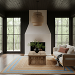 A styled living room interior with a white fireplace wall, dark wood ceiling and trim, large black-framed windows looking out to trees, a woven pendant light, a wooden coffee table, a light sofa with pillows, and a patterned area rug.