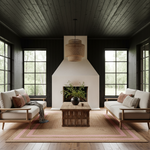 A styled living room interior with two cream upholstered armchairs facing a low rattan coffee table, set on a large woven area rug with pink border stripes. The room features a white chimney-style fireplace, dark wood-paneled walls and ceiling, large black-framed windows, and a green plant in a dark vase.