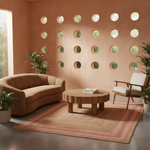A styled living room scene with a curved tan sofa, a round wooden coffee table, a pink-and-tan patterned rug, a mid-century armchair, and potted plants against a terracotta wall with circular cutouts.