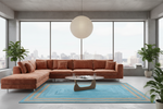A modern living room featuring a large rust-brown sectional sofa, a bright blue patterned area rug, a round glass-top coffee table, and a large white paper lantern pendant light, set against a concrete-walled window backdrop with a city skyline.