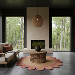 A styled interior living room scene with a round woven rattan pendant light hanging above a low round coffee table on a scalloped jute rug. The room has dark wood walls and ceiling, large windows showing a forest view, a beige sofa with patterned pillows, and a central fireplace opening.