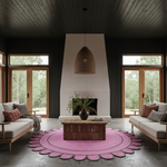 A modern living room with a large pink scalloped area rug under a wooden coffee table, flanked by two light gray sofas with wood frames and dark pillows. The room features a tall white fireplace surround, dark wood-paneled ceiling, and large windows looking out to green trees.