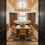 A modern dining room with a wooden dining table and six woven-back chairs, set on a floral patterned rug. The room features a light wood ceiling, recessed lighting, and a contemporary kitchen in the background, viewed through open dark wood doors.