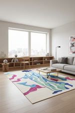 A modern living room featuring a large cream-colored area rug with abstract wavy patterns in blue, green, pink, and purple. The room includes a light gray sofa, a wooden coffee table, a built-in wooden bench by large windows, and light wood flooring.