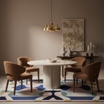 A styled dining room scene with a round white stone dining table surrounded by four upholstered brown chairs on a colorful abstract area rug. A brass pendant light hangs above, and a textured wall art piece and side console are visible in the background.
